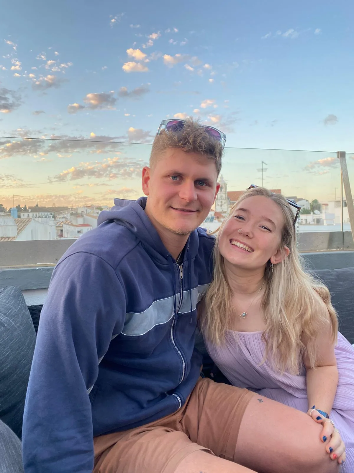 Tessa and Callum on a rooftop terrace at sunset with a city skyline behind them.