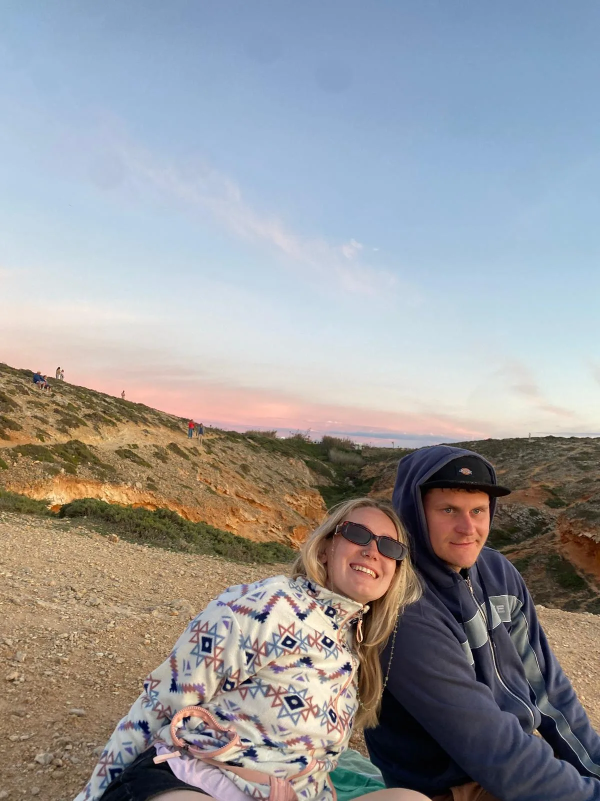 Tessa and Callum sitting on a clifftop above the sea as the sky turns pink.