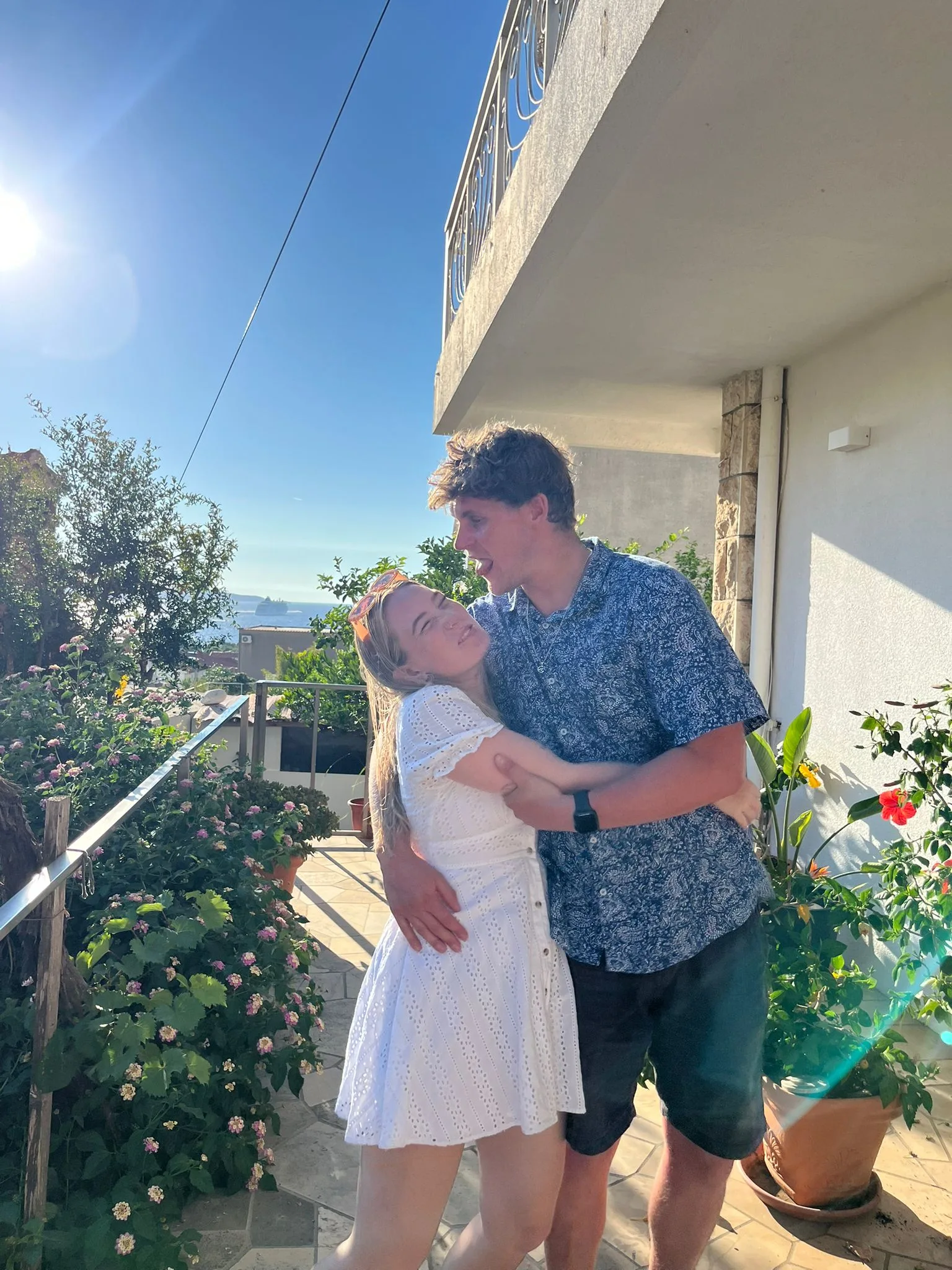 Callum hugging Tessa on a sunlit terrace surrounded by flowers.
