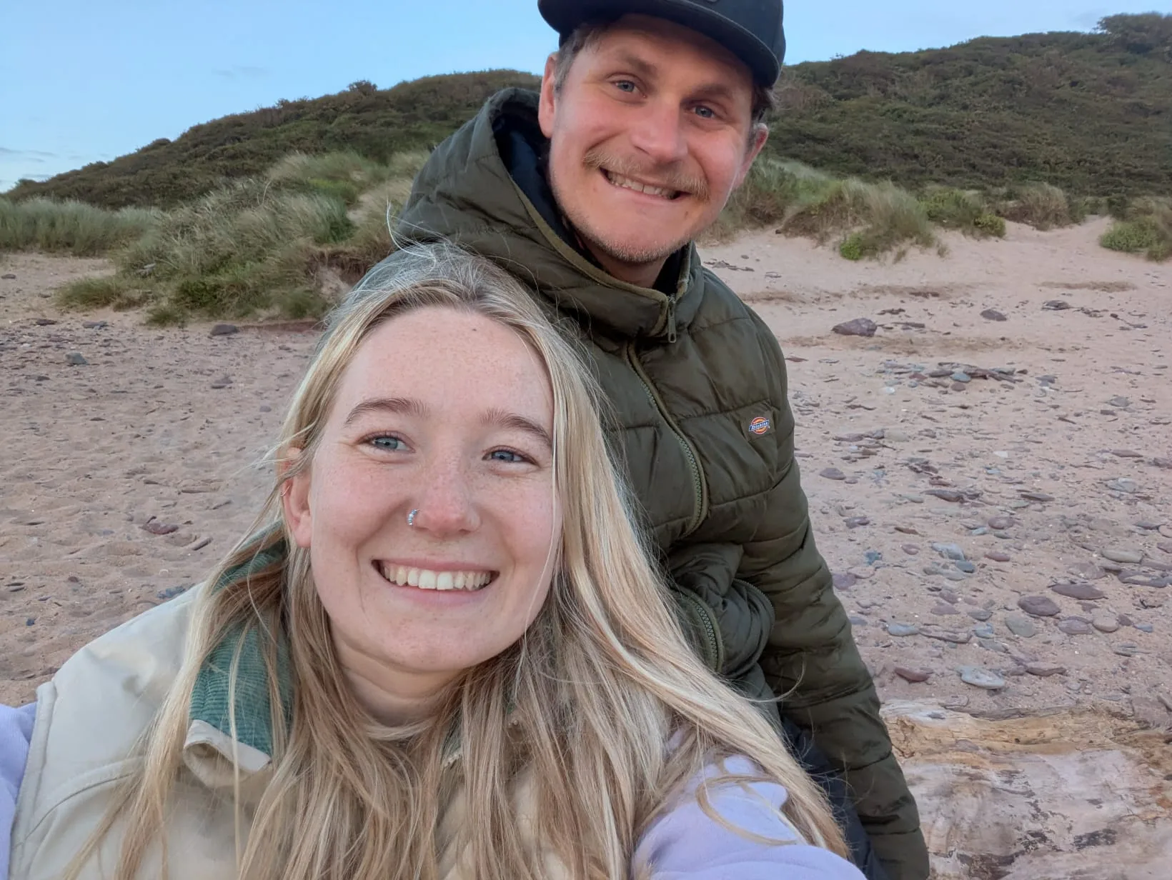 Tessa and Callum smiling on a sandy beach among the dunes at dusk.