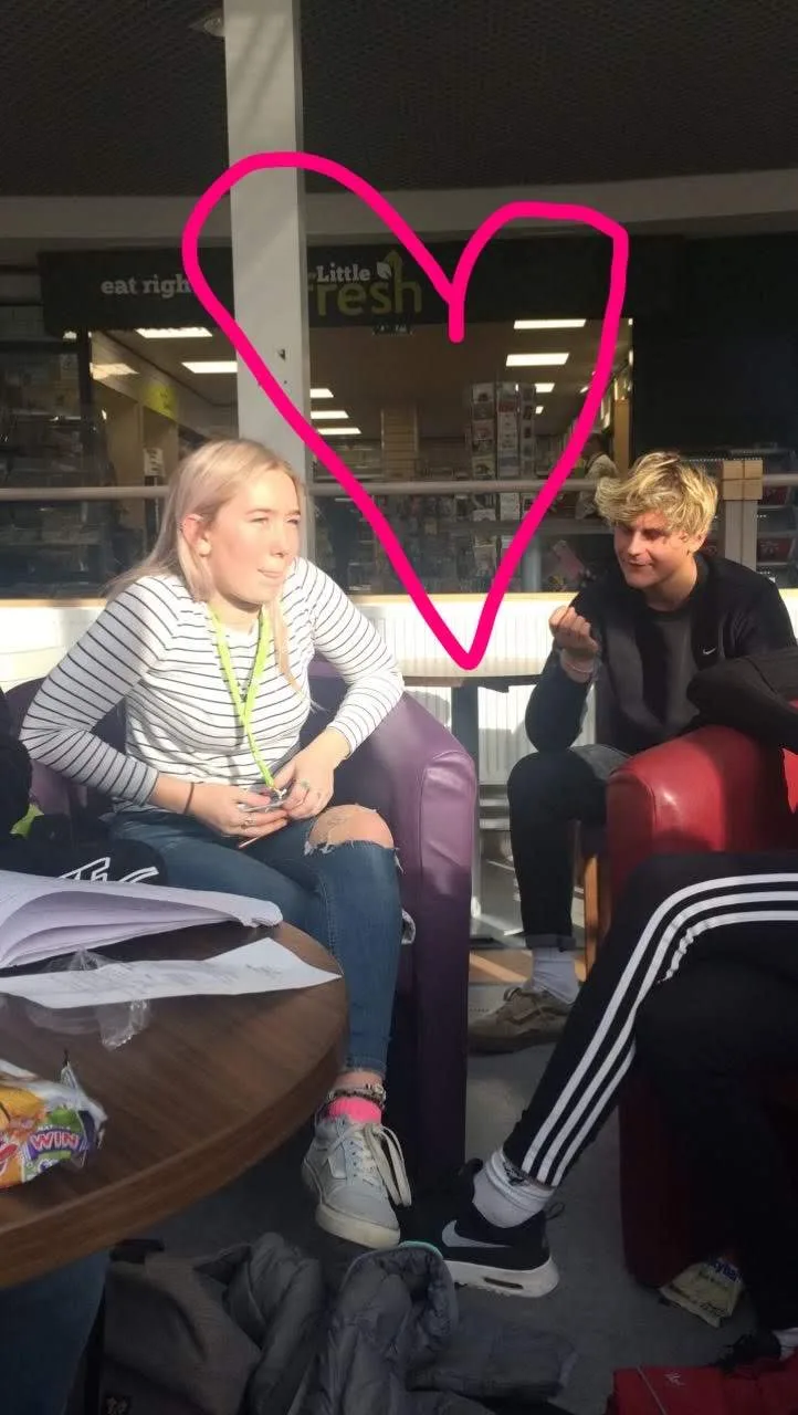 Tessa and Callum sitting together in a college common room, with a pink heart doodled over the photo.